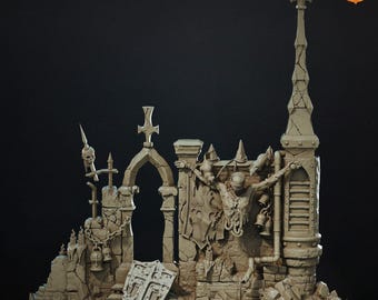 Puede incluir: Un diorama en miniatura de color beige que presenta un muro de piedra en ruinas con una aguja alta, un arco apuntado y una figura central atada a la estructura. La escena incluye un escudo con un emblema de cruz, campanas colgantes y cadenas.