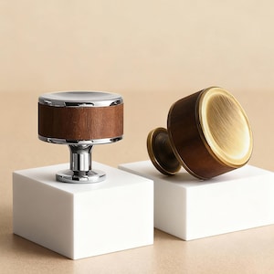 Puede incluir: Dos tiradores decorativos para armarios. Uno presenta una parte superior y una base plateadas con una banda de madera. El otro tiene un acabado de latón y un cuerpo de madera. Ambos se muestran sobre bloques blancos sobre un fondo neutro.