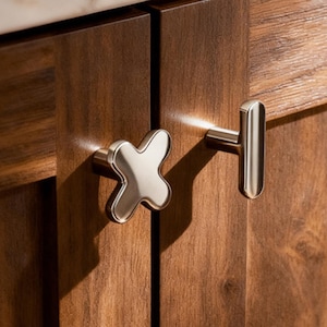 May include: Close-up of two cabinet doors with decorative hardware. One door has a silver cross-shaped knob, and the other has a silver T-shaped handle. The doors are made of dark brown wood, and a white marble countertop is visible.