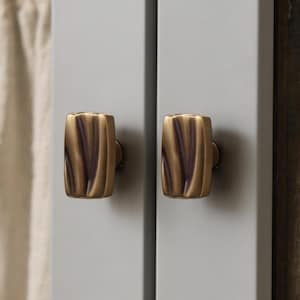 Könnte beinhalten: Nahaufnahme von zwei bronzefarbenen Schrankknöpfen mit einem strukturierten, vertikalen Rillendesign. Die Knöpfe sind an einer hellgrauen Schranktür befestigt. Beige Vorhänge sind im Hintergrund zu sehen.