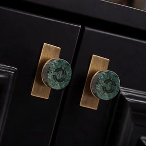 May include: Two circular cabinet knobs featuring a green marble face with a black circular logo inlay, mounted on a rectangular brushed brass backplate. The hardware is attached to a black cabinet door with a recessed panel design and visible edges.