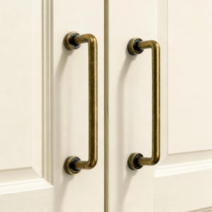 Puede incluir: Dos tiradores de armario de latón antiguo con un diseño rectangular curvo. Los tiradores están montados en una puerta de armario de color crema. El herraje tiene un aspecto vintage y envejecido, añadiendo un toque de elegancia clásica.