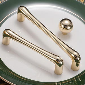 May include: Gold-toned cabinet hardware set, including two pull handles and a round knob. The hardware is displayed on a white plate with a green and gold border, showcasing a modern design.