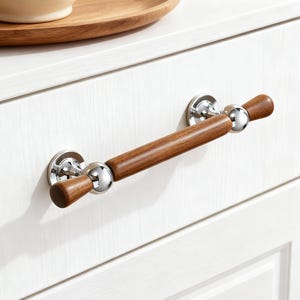 May include: A cabinet pull with a polished chrome finish and a dark brown wooden handle. The handle is cylindrical with rounded ends. The pull is mounted on a white cabinet door.