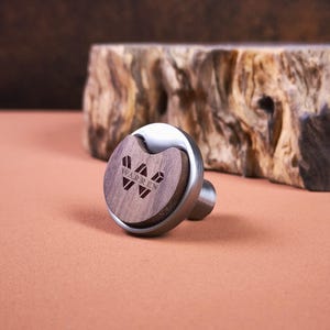 Puede incluir: Un pomo circular para muebles con una base de metal plateado cepillado y una incrustación de madera marrón oscura. El centro de madera está grabado con el texto WARREN y un logotipo geométrico en forma de W. El pomo tiene un perfil redondeado.