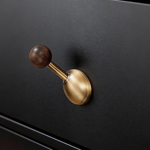 Puede incluir: Primer plano de un tirador de cajón. El tirador presenta una esfera de madera oscura sobre un vástago de latón y una base de latón. El tirador está montado en un cajón negro, con el herraje ligeramente inclinado hacia afuera. El herraje tiene una estética vintage.