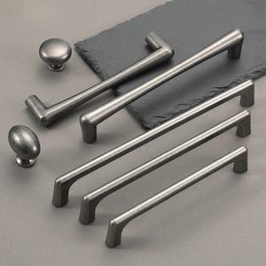 May include: A selection of antique-style cabinet hardware, comprising two oval knobs, two round knobs, and four bar pulls. The hardware is a muted silver colour, with a textured finish. The pieces are arranged on a dark grey surface.