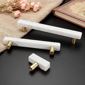 May include: Three white marble cabinet pulls with brass hardware. Two are long, rectangular pulls, and one is a shorter, square pull. The pulls are arranged on a dark surface, with a neutral-colored fabric and flowers in the background.