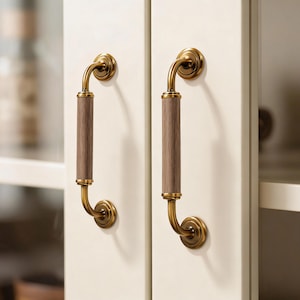 May include: Two cabinet handles with a brass finish and wooden center. The handles are mounted on a cream-colored cabinet door. The wooden center has a light brown color.