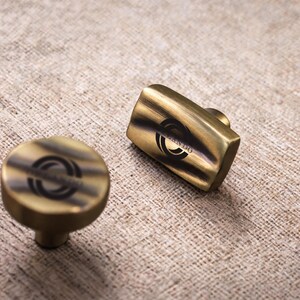May include: Two brass-toned metal cabinet knobs with a brushed finish. One knob is circular and the other is rectangular with rounded edges. Both feature a black circular logo with the text "ORLANDO" printed in the centre of the top surface.