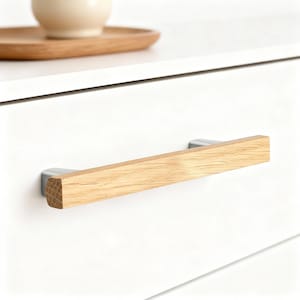 May include: A close-up of a white cabinet drawer with a wooden handle. The handle is rectangular, made of light-colored wood, and attached to the drawer with silver-colored metal brackets. The wood grain is visible. A wooden tray and a beige ceramic object are visible in the background.
