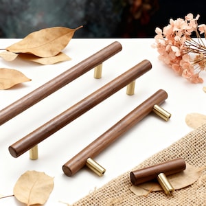 May include: A collection of wooden cabinet pulls with brass-colored hardware. The pulls are a rich brown color and are arranged on a white surface with dried leaves and flowers.