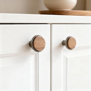 May include: Close-up of two cabinet knobs. Each knob features a round, light-colored wood center set within a dark metal ring. The knobs are attached to a white cabinet door. A wooden tray and a white bowl are visible in the background.