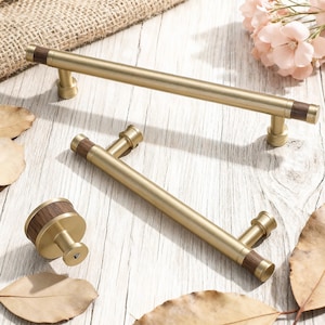 May include: A set of gold-toned cabinet hardware, including two bar pulls and a knob. The pulls have a cylindrical design with dark wood accents on the ends. The knob features a matching wood and gold finish. The hardware is displayed on a light wood surface.
