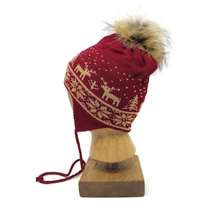 Pode incluir: Um gorro de inverno de malha vermelha com um padrão de renas e flocos de neve em bege. O gorro tem protetores de orelha, uma fita para o queixo e um pompom marrom fofo. O gorro é exibido em uma cabeça de madeira.