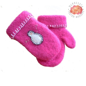 Peut inclure: Une paire de moufles en laine rose vif avec un appliqué de bonhomme de neige blanc. Les moufles ont des coutures blanches autour des poignets et du bonhomme de neige. L'image comprend un logo "Minnesota Custom Woolens".