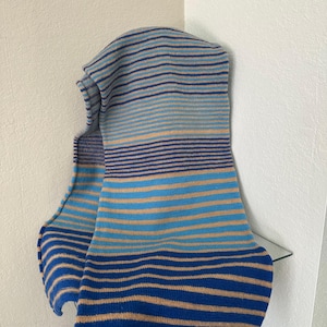 May include: A knitted scarf featuring horizontal stripes in shades of tan, light blue, and dark blue. The scarf has a consistent ribbed texture throughout, with varying widths of color bands that create a repeating pattern across the entire length.