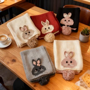May include: A collection of knitted scarves in various colors, including cream, red, black, and gray. Each scarf features an embroidered bunny face and a matching pom-pom. The scarves are displayed on a wooden table.
