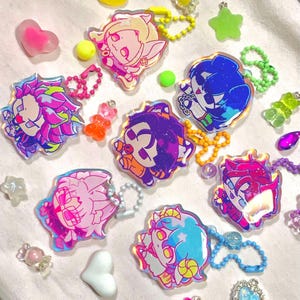 Könnte beinhalten: Eine Sammlung farbenfroher Schlüsselanhänger im Anime-Stil mit schillernden Akzenten. Jeder Schlüsselanhänger zeigt ein einzigartiges Charakterdesign mit leuchtenden Farben wie Pink, Blau und Lila. Verschiedene kleine, bunte Süßigkeiten und Charms umgeben die Schlüsselanhänger.