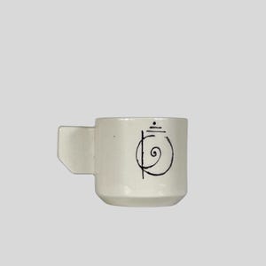 May include: A small, off-white ceramic mug with a square handle. The mug features a black, stylized design on the front. The mug is designed for hot beverages.