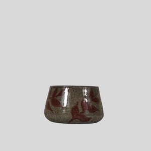 Puede incluir: Un cuenco de cerámica pequeño y redondo con un esmalte moteado en tonos tierra. El cuenco está adornado con un patrón de formas de hojas de color rojo oscuro. El diseño es sencillo y elegante, adecuado para servir o exhibir.