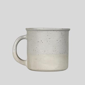 May include: A two-tone ceramic mug with a rounded handle. The top half is a speckled grey, and the bottom half is a creamy white. The mug has a simple, classic design, suitable for coffee or tea.