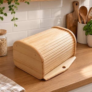 May include: A light-colored wooden bread box featuring a curved, slatted roll-top lid that slides open. The rectangular container has rounded side panels and a flat base with a small circular cutout at the front edge for easy opening and closing.