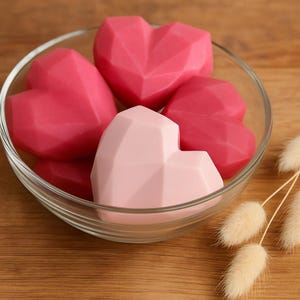 May include: A clear glass bowl filled with geometric heart-shaped objects. The hearts are in shades of pink, with a lighter pink heart in the foreground. The hearts have a faceted design. Dried flowers are in the background.