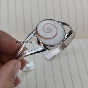 May include: A silver cuff bracelet with a large, round, white stone displaying a brown spiral design. The bracelet has a simple, open design. The brand name "NAVOJEWELCO" is visible on the side of the bracelet.