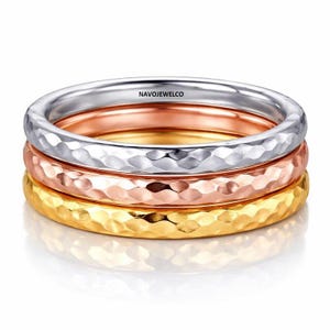 May include: Three stackable rings in silver, rose gold, and gold. Each ring has a hammered texture. The rings are displayed on a white surface with a slight reflection. The brand name "NAVOJEWELCO" is visible on the top ring.