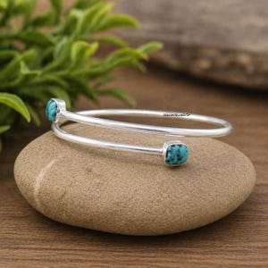 May include: A silver-colored cuff bracelet with two square turquoise stones. The bracelet has a minimalist design and is resting on a smooth, light brown stone. The background includes green foliage and a wooden surface.