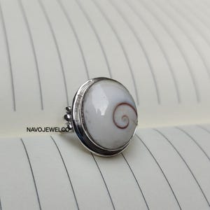 Puede incluir: Anillo de plata con una piedra blanca redonda y grande con un diseño en espiral marrón. El anillo tiene una banda sencilla y un engaste decorativo. La marca "NAVOJEWELCO" es visible en el lateral.