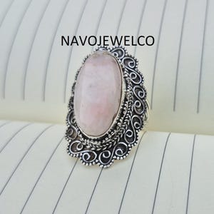 May include: A silver ring with a large, oval, pale pink gemstone. The gemstone is set in a decorative, filigree-style setting. The ring is displayed on a lined surface, with the text "NAVOJEWELCO" visible.