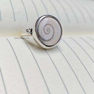 May include: A silver ring with a large, round, off-white stone featuring a spiral design. The ring has a simple band and a decorative element near the stone. The text "NAVOJEWELCO" is visible on the band.