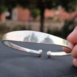 Puede incluir: Un brazalete de puño abierto de color plateado. El brazalete tiene un diseño liso y curvado con extremos redondeados. El metal refleja la luz y el brazalete parece estar hecho de un material sólido.