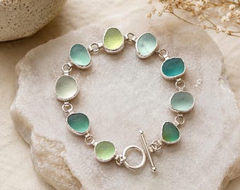 May include: A bracelet featuring smooth, rounded pieces of sea glass in various shades of green and teal, connected by silver metal links. The piece is secured with a silver toggle clasp, creating a circular arrangement of polished glass elements.