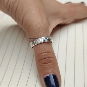Puede incluir: Un anillo de plata con un diseño de patrón de ondas repetidas. El anillo se lleva en un dedo, mostrando los intrincados detalles de la joya. El anillo es una banda simple con un acabado pulido.