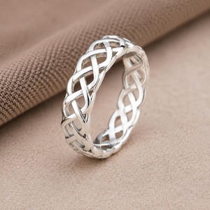 Peut inclure: Une bague en argent poli présentant un motif complexe de nœuds tressés continus qui fait le tour de l'anneau. La surface métallique est lisse et réfléchissante, mettant en valeur un design ajouré détaillé formant des boucles entrelacées.