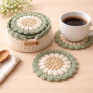 May include: A crocheted tea set with a matching container, two coasters, and a cup of coffee. The set features a floral design in cream, tan, and green. The container has a wooden tag that reads "mandiadia".