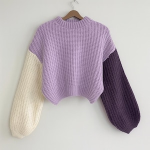 May include: A lilac-coloured, chunky knit sweater with a round neckline and cream and dark purple sleeves. The sweater is hanging on a wooden hanger against a white background.