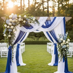 May include: A wooden wedding arch adorned with flowing white and royal blue fabric. The arch is decorated with white roses, blue hydrangeas, and foliage. White chairs are arranged on the green grass, creating a picturesque outdoor wedding setting.