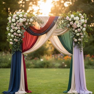 May include: A wedding arch decorated with white and pink roses, greenery, and flowing fabric in shades of red, navy, cream, green, and lavender. The arch is set on a green lawn with sunlight in the background.