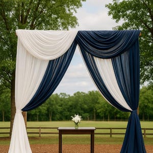 Peut inclure: Une arche de mariage drapée de tissu blanc et bleu marine. Le tissu est disposé en plis fluides, créant une toile de fond douce et élégante. Une petite table en bois avec un bouquet de fleurs blanches est placée devant l'arche, en extérieur.