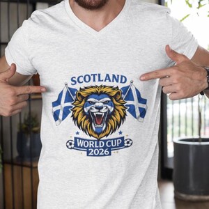 May include: A white V-neck t-shirt featuring a graphic of a lion with the Scotland flag colors. The text "SCOTLAND" is above the lion, and "WORLD CUP 2026" is below. Two flags with the Scotland flag design are on either side of the lion.