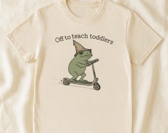 May include: Cream-colored t-shirt featuring a whimsical illustration of a green frog wearing a hat and riding a scooter. The text "Off to teach toddlers" is printed above the frog. The shirt has short sleeves and a crew neck.