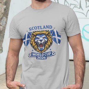 May include: A light gray t-shirt featuring a lion's head with a blue and white Scottish flag design. The text "SCOTLAND" is above the lion, and "WORLD CUP 2026" is below. Two Scottish flags flank the lion.