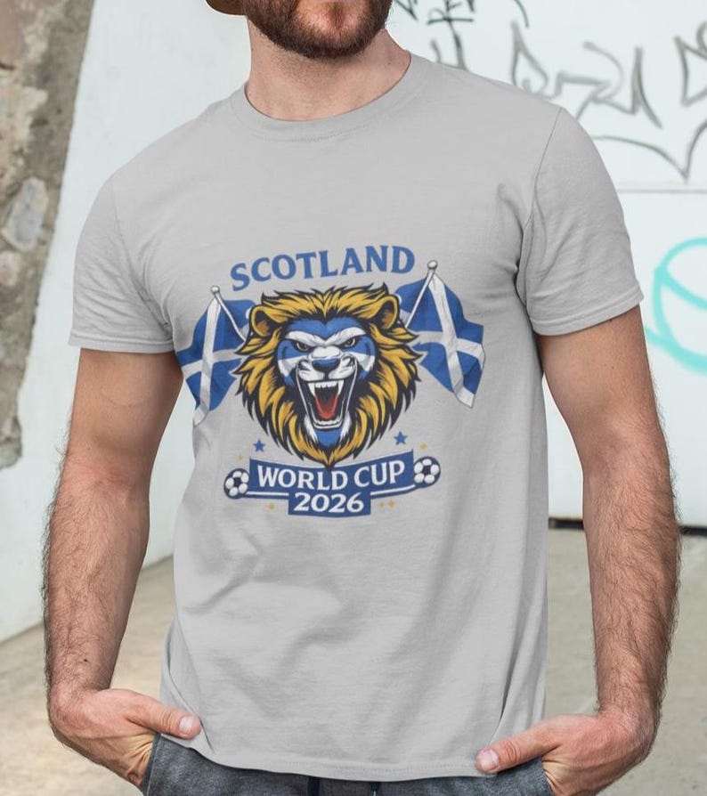 May include: A light gray t-shirt featuring a lion's head with a blue and white Scottish flag design. The text "SCOTLAND" is above the lion, and "WORLD CUP 2026" is below. Two Scottish flags flank the lion.
