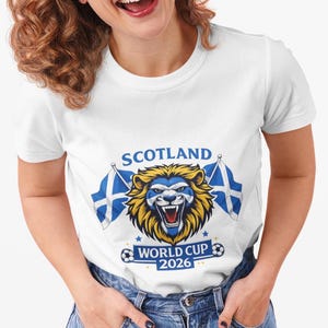 May include: White t-shirt featuring a lion graphic with the text "SCOTLAND WORLD CUP 2026". The lion has a blue and yellow design, with two Scottish flags on either side. The shirt is a crew neck style.