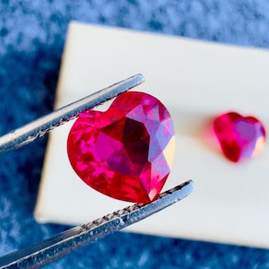 May include: A close-up of two heart-shaped, faceted, red gemstones. One is held by silver tweezers, the other rests on a white surface. The gemstones are a vibrant shade of red, with multiple facets catching the light.