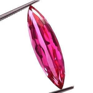 May include: A marquise-cut gemstone, held by tweezers. The gem is a deep pink colour, with multiple facets that reflect light. The tweezers are silver-coloured. The background is plain white.
